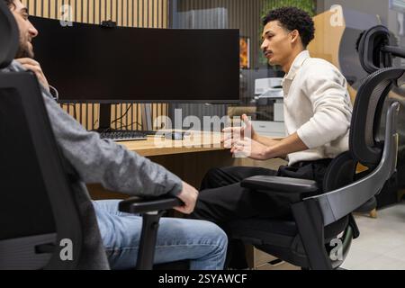 Two colleagues in a modern office engage in a focused discussion. A sleek computer setup and comfortable chairs create an ideal environment for produc Stock Photo