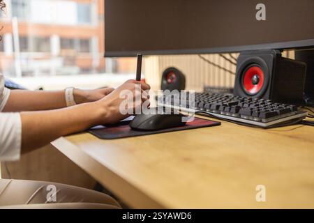A person works at a modern office desk with a computer setup. The focus is on the hands using a digital tablet and stylus, surrounded by keyboard, mou Stock Photo