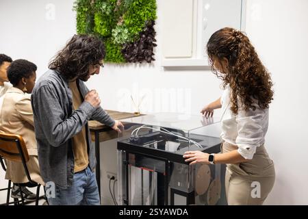 A diverse team collaborates in a modern office. Colleagues engage with a 3D printer while others focus on tasks, creating an atmosphere of innovation Stock Photo