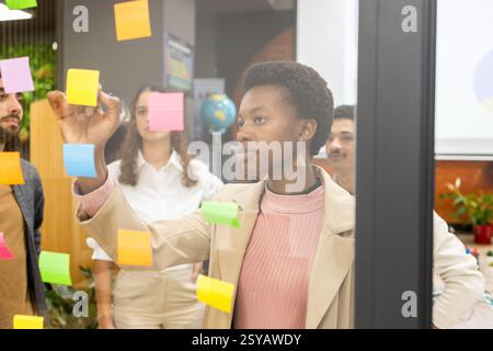 A diverse office team collaborates during a brainstorming session, using colorful sticky notes on a glass wall. The atmosphere is creative and focused Stock Photo