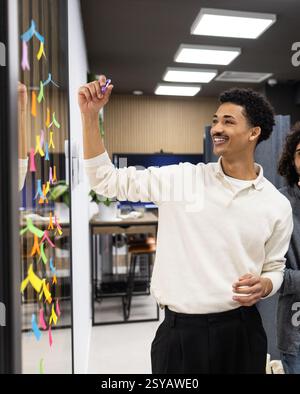 A professional in a modern office setting adds colorful sticky notes to a glass wall during a brainstorming session. The image captures teamwork and c Stock Photo