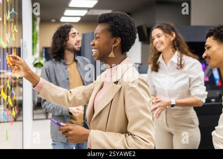 A diverse group of colleagues engaged in a brainstorming session in a modern office. Smiling and interacting, they are using colorful sticky notes on Stock Photo