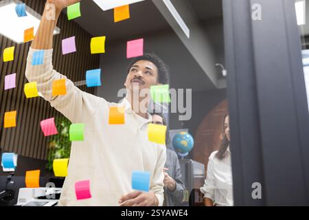 A group of professionals brainstorms using vibrant sticky notes on glass in a modern office. This teamwork fosters creativity, collaboration, and idea Stock Photo
