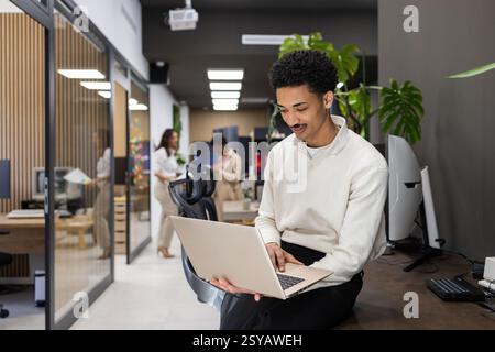 A professional office environment with a man using a laptop while sitting on a desk, and colleagues collaborating in the background, surrounded by mod Stock Photo