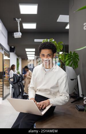 A cheerful professional sits on a desk in an open, modern office environment, working on a laptop. Colleagues in the background contribute to a collab Stock Photo