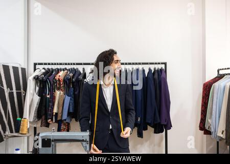 A tailor in a navy suit smiles while holding a measuring tape in a suit store. Behind him, neatly arranged jackets on hangers create a stylish backdro Stock Photo