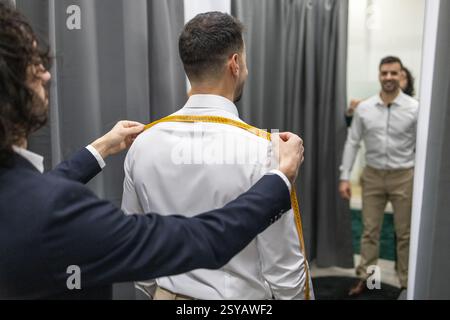 A professional tailor measures a client shoulder width using a tape measure in a fitting room. Both are well dressed, highlighting the tailoring proce Stock Photo