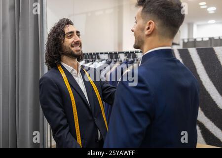 A tailor, measuring tape around his neck, interacts with a client during a suit fitting in a suit store, showcasing professionalism and personal servi Stock Photo