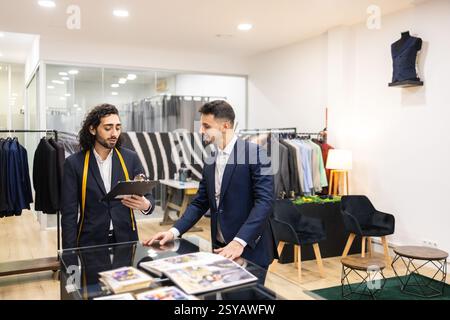 A tailor discusses suit customization with a client in a suit store. The space is stylish, with suits displayed and a mannequin visible. Professional Stock Photo