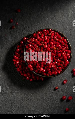 Top view of a dark bowl overflowing with bright red peppercorns, set against a contrasting textured dark background. The vibrant color of the pepperco Stock Photo