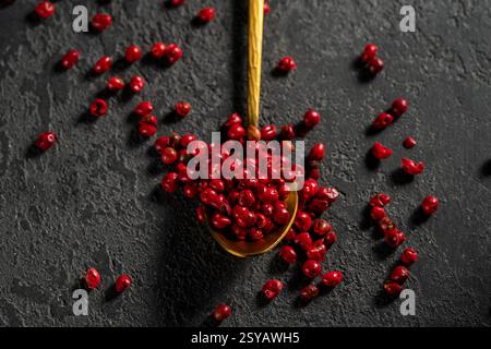 Top view of bright red peppercorns scattered from a golden spoon against a textured dark background, showcasing vivid color and texture contrasts in a Stock Photo