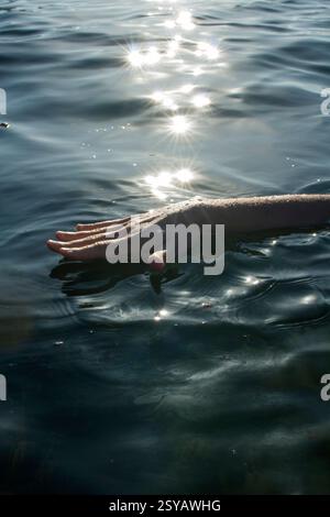 A hand gently touches the surface of a tranquil water body, creating ripples. Sunlight sparkles on the water, enhancing the peaceful atmosphere of the Stock Photo