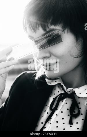 Black and white portrait of a woman holding a film strip over her eyes, evoking a sense of mystery and nostalgia, with sunlight in the background. Stock Photo