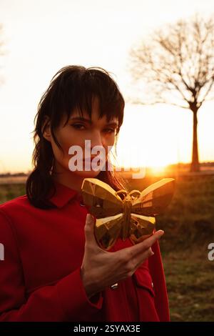 A Middle Eastern woman stands outside during sunset, holding a golden butterfly. The soft light highlights her face, creating a serene and ethereal at Stock Photo