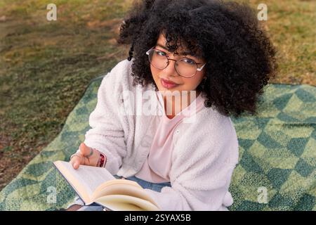A young latin woman with curly hair and glasses sits on a picnic blanket outdoors, absorbed in reading a book. The serene setting suggests relaxation Stock Photo