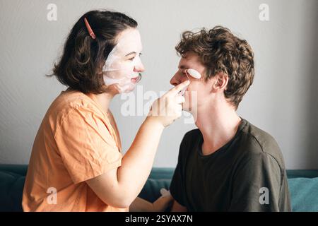A young man and woman enjoy a playful skincare routine together in their living room. Both are smiling gently, she is covered in face masks, using a p Stock Photo