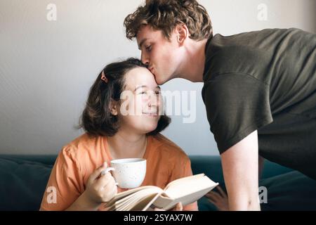 A man affectionately kissing a woman's forehead while she enjoys reading a book and drinking from a mug, comfortably resting at home. Stock Photo