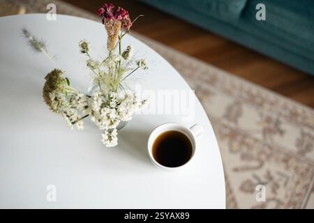 Top view of a serene home scene featuring a cup of black coffee and a vase of delicate dried flowers on a sleek white table, evoking a peaceful, restf Stock Photo