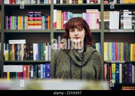 A young woman stands confidently in front of a vibrant, well-stocked bookcase, her expression thoughtful and serene. Stock Photo