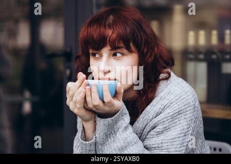 A young woman with red hair sips from a blue cup of coffee, wrapped in a cozy sweater. She gazes thoughtfully, creating a peaceful and introspective a Stock Photo