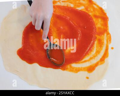 A child spreads rich tomato sauce evenly over fresh pizza dough with a spoon, preparing for toppings. A vibrant scene capturing a homemade pizza makin Stock Photo
