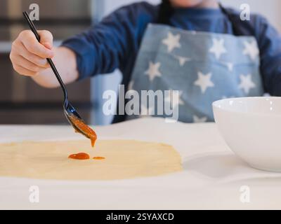 A child prepares homemade pizza, spreading tomato sauce on a rolled out dough using a spoon. They wear an apron with stars, highlighting a cozy cookin Stock Photo