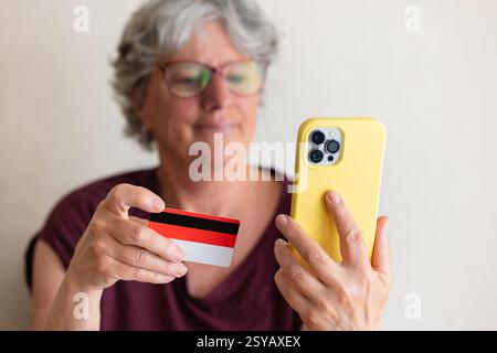 A senior woman with gray hair uses a smartphone and holds a credit card. She is smiling, suggesting ease and confidence in online shopping or banking. Stock Photo