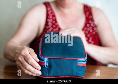 A senior woman sits at a table holding a digital device. The focus is on the bag and their hands, with a blurred background showing a polka dot dress. Stock Photo