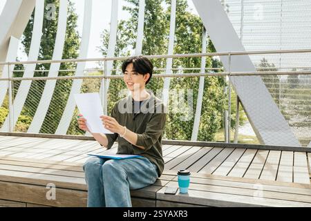 A young man study outside on wooden steps. The student smiling and reading notes. The atmosphere is casual, ideal for motivation and study. Stock Photo