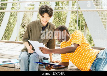 Two friends sit on wooden steps outdoors, discussing documents. One, in a striped shirt, leans in to listen attentively. Their expressions show eagern Stock Photo