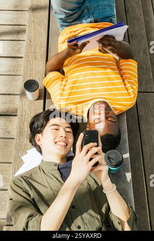 Two friends enjoy a sunny day outdoors. One is lying back with a notebook, while the other smiles at a smartphone. A casual scene of friendship and le Stock Photo