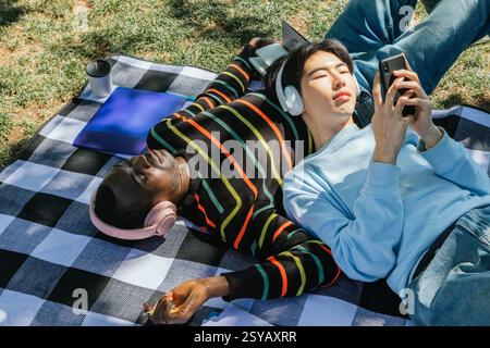 Two friends enjoy a sunny day, lying on a checkered blanket. One listens to music with headphones, eyes closed, while the other uses a smartphone. Nea Stock Photo