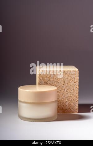 An elegant cosmetic cream jar paired with a natural sponge, both displayed on a minimalist stone podium against a soft gray background, ideal for beau Stock Photo