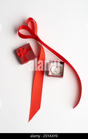 From above, a romantic Valentine's Day setup featuring a red ribbon arranged in a heart shape, with a small red gift box and a ring box containing two Stock Photo