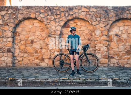 A young male cyclist stands next to his road bike, leaning against an aged stone wall in Zahara de los Atunes, Cadiz. The man is wearing sunglasses, a Stock Photo