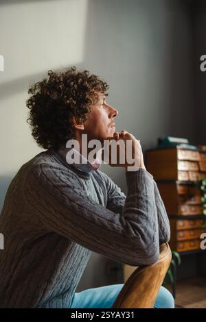A contemplative man with curly hair sits by a window, resting his chin on his hands. Warm natural light highlights his face, creating a peaceful and i Stock Photo