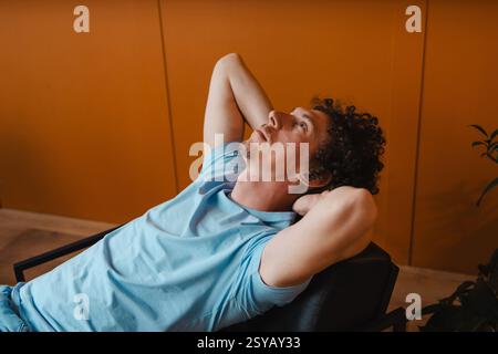 A curly-haired man leans back in a chair with his hands behind his head, gazing upward. The warm orange wall and casual blue t-shirt create a relaxed, Stock Photo