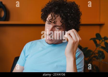 A young man with curly hair in a light blue shirt, sitting thoughtfully in a room with an orange background and green plants. The scene conveys a casu Stock Photo
