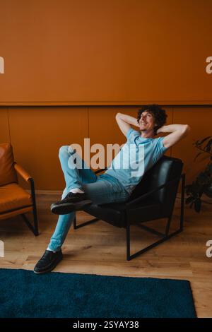 A young man with curly hair relaxes comfortably in a modern chair. His casual attire complements the stylish interior setting, creating a laid-back at Stock Photo
