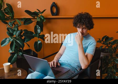 A curly-haired young man in a light blue shirt works on his laptop in a stylish room with plants and a warm orange wall, looking thoughtful and focuse Stock Photo