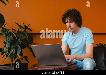 A curly-haired young man focused on his laptop, sitting in a modern space with orange walls and greenery. Casual attire, relaxed atmosphere, productiv Stock Photo