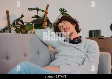 A curly-haired young man with casual attire, relaxing on a sofa with headphones around his neck. The serene setting features indoor plants in the back Stock Photo