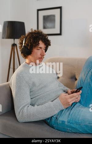 A young man with curly hair relaxes on a sofa, wearing a gray sweater, blue jeans, and headphones. He is focused on his phone, creating a calm atmosph Stock Photo