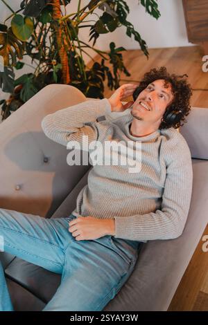 Young man with curly hair relaxes on a modern gray couch, wearing headphones and enjoying music. He has a cozy sweater, blue jeans, and a content expr Stock Photo