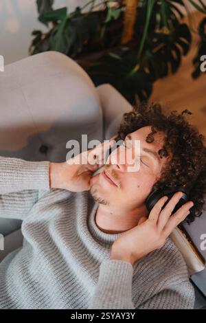 A young man with curly hair relaxes in an armchair, eyes closed, listening to music on headphones. He wears a cozy gray sweater, surrounded by lush gr Stock Photo