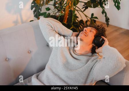 A young man with curly hair relaxes on a sofa, listening to music with headphones. Sunlight streams through a window, plants in the background, creati Stock Photo