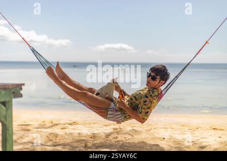 A woman lounges in a colorful hammock on a sandy beach in the Philippines, sharing a playful moment with a small cat under a clear blue sky. Stock Photo