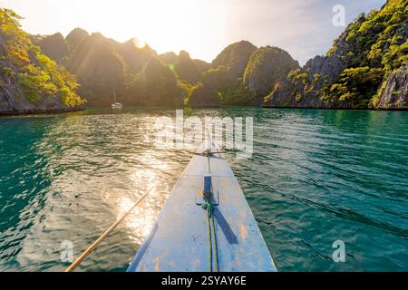 A view from a boat gliding over calm waters surrounded by lush, rocky mountains in the Philippines, bathed in the glow of the setting sun. Stock Photo