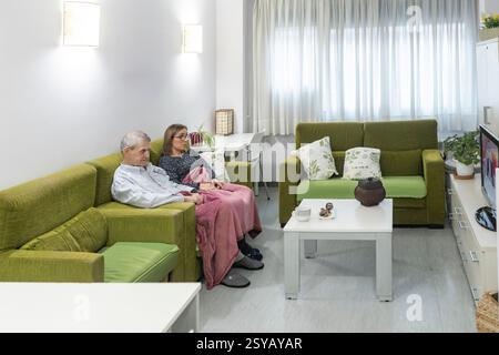 Couple with intellectual disabilities sitting comfortably on a green couch in a supervised home, watching television. The room is decorated with plant Stock Photo