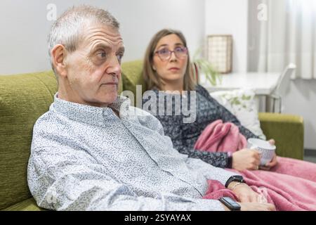 Couple with intellectual disabilities sitting on a green couch at supervised home, wrapped in a pink blanket. They are relaxed, with a cup of coffee, Stock Photo